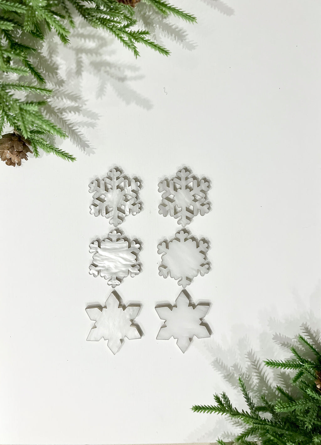 Six white translucent snowflake-shaped cutouts arranged in two columns on a white background with pine branches and cones