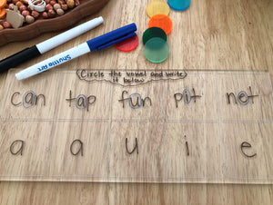 Transparent board with words containing vowels written inside boxes, colorful translucent discs, and two dry erase markers on wood surface