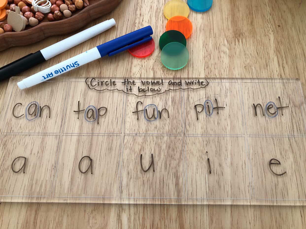 Transparent board with words containing vowels written inside boxes, colorful translucent discs, and two dry erase markers on wood surface