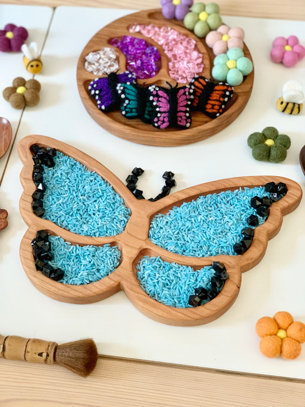Wooden butterfly-shaped sensory tray filled with blue rice and black gem accents on white surface with felt flowers and bees