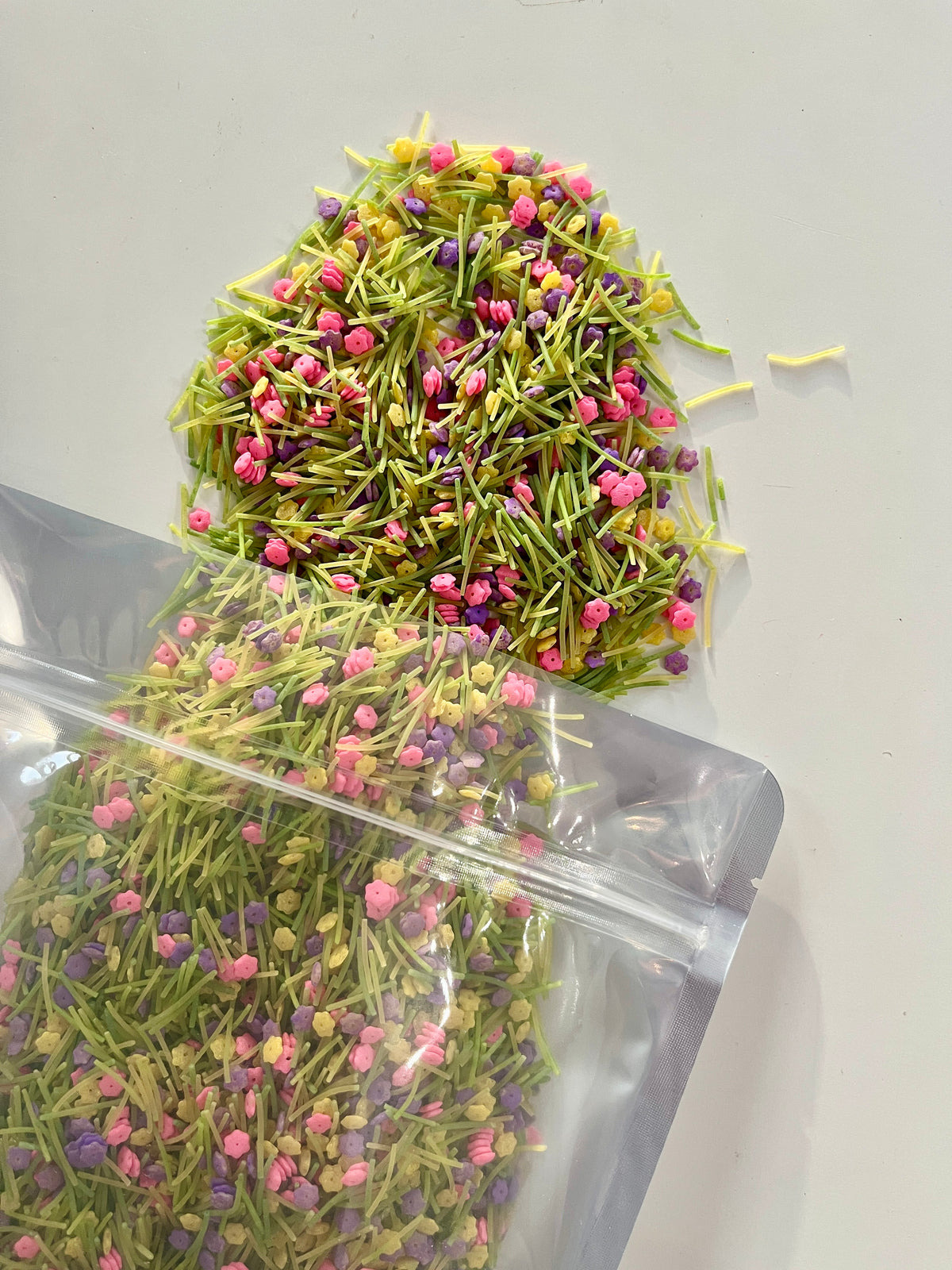 Clear resealable pouch spilling thin green pasta strands mixed with small pink and purple flower-shaped sprinkles on white background