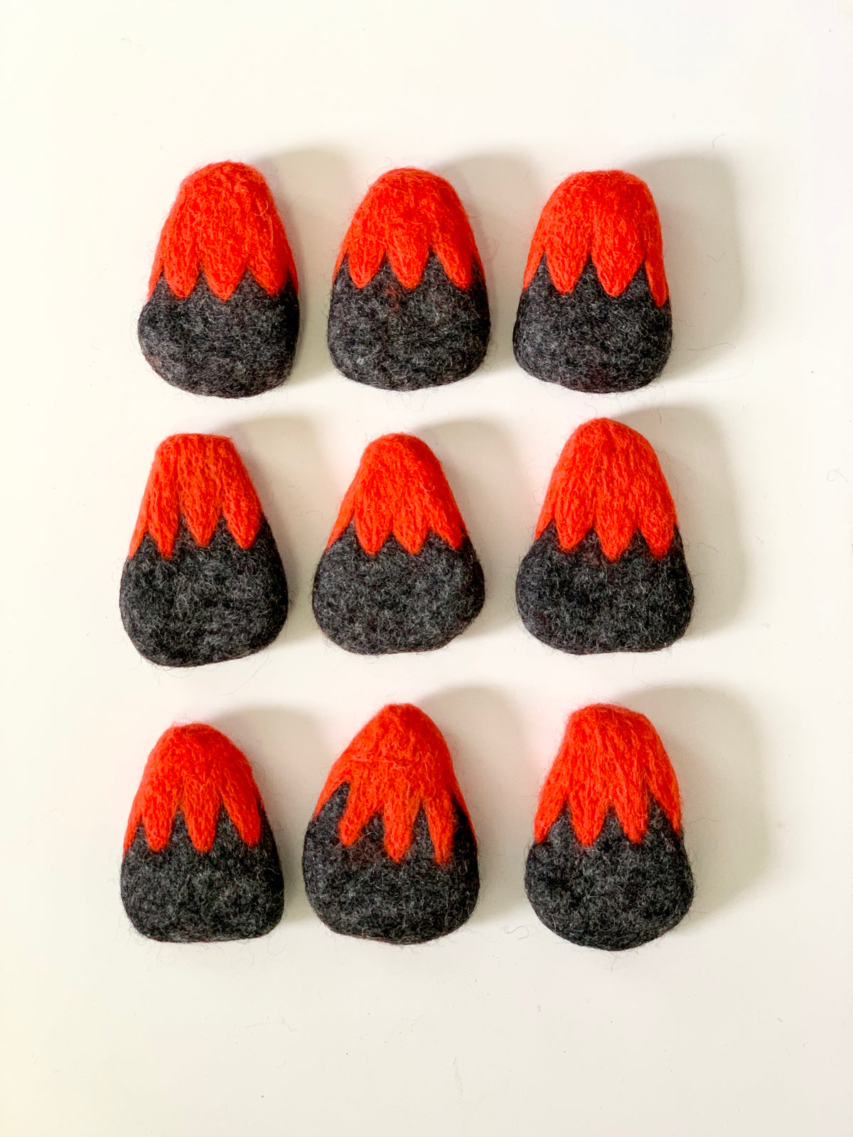 Nine small handmade felt volcanoes with red tops and dark gray bases arranged on white background