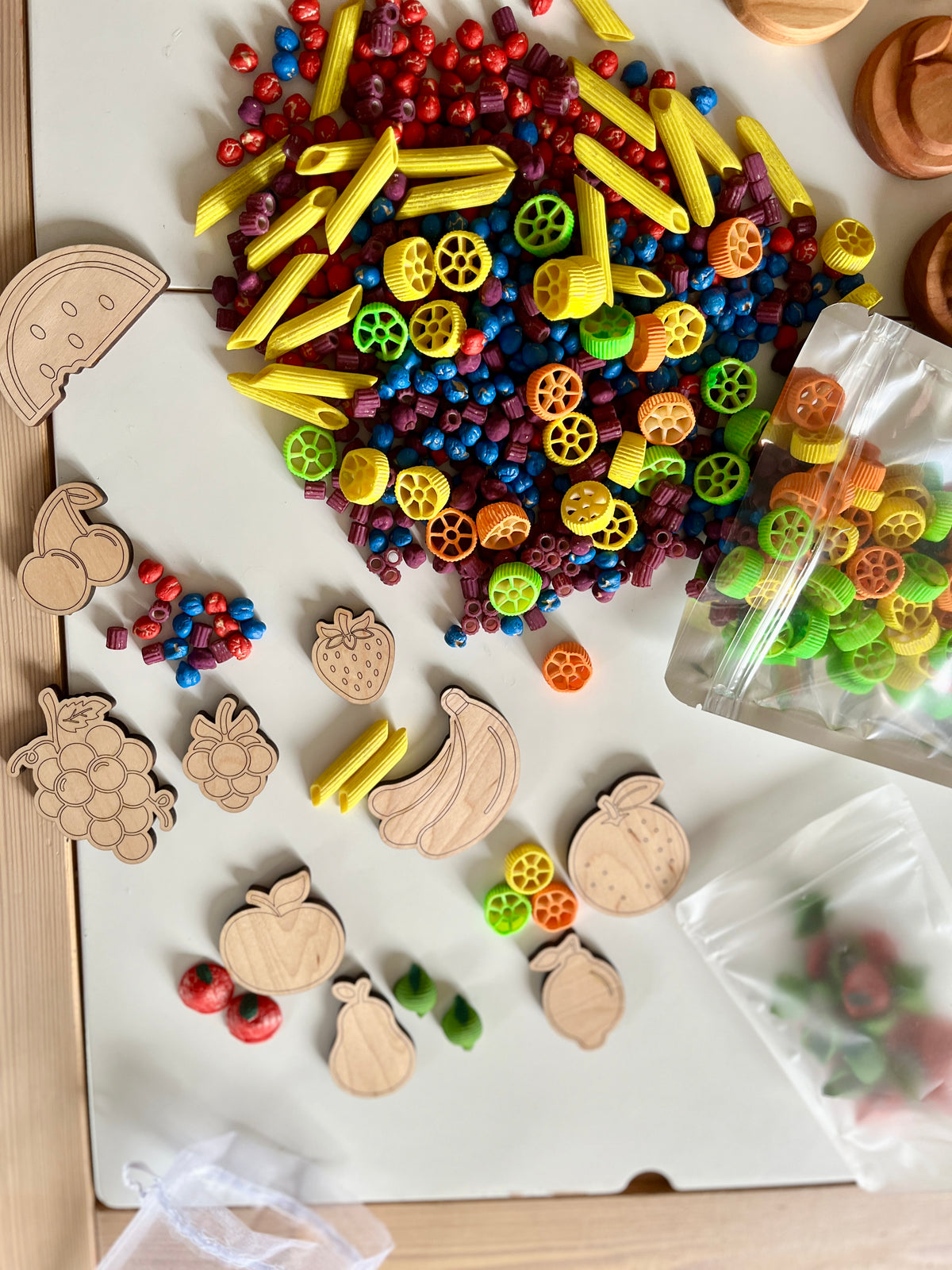 Colorful pasta shapes and dried beads mixed with wooden fruit cutouts on a white surface with small bags nearby