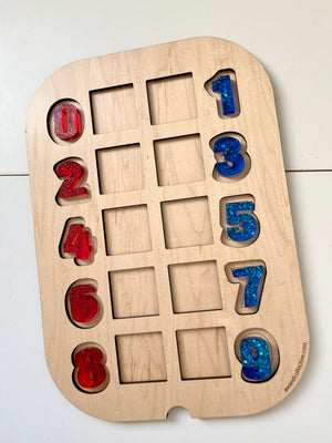 Red and blue glitter-filled resin numbers arranged on a light wood puzzle board with empty slots on a white surface