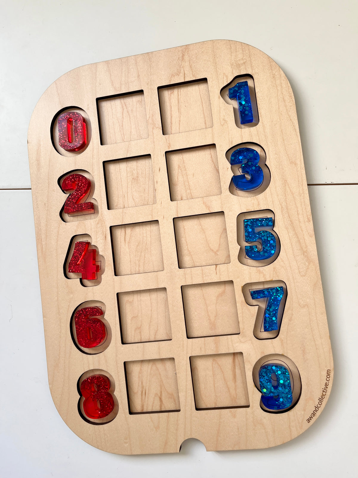Red and blue glitter-filled resin numbers arranged on a light wood puzzle board with empty slots on a white surface