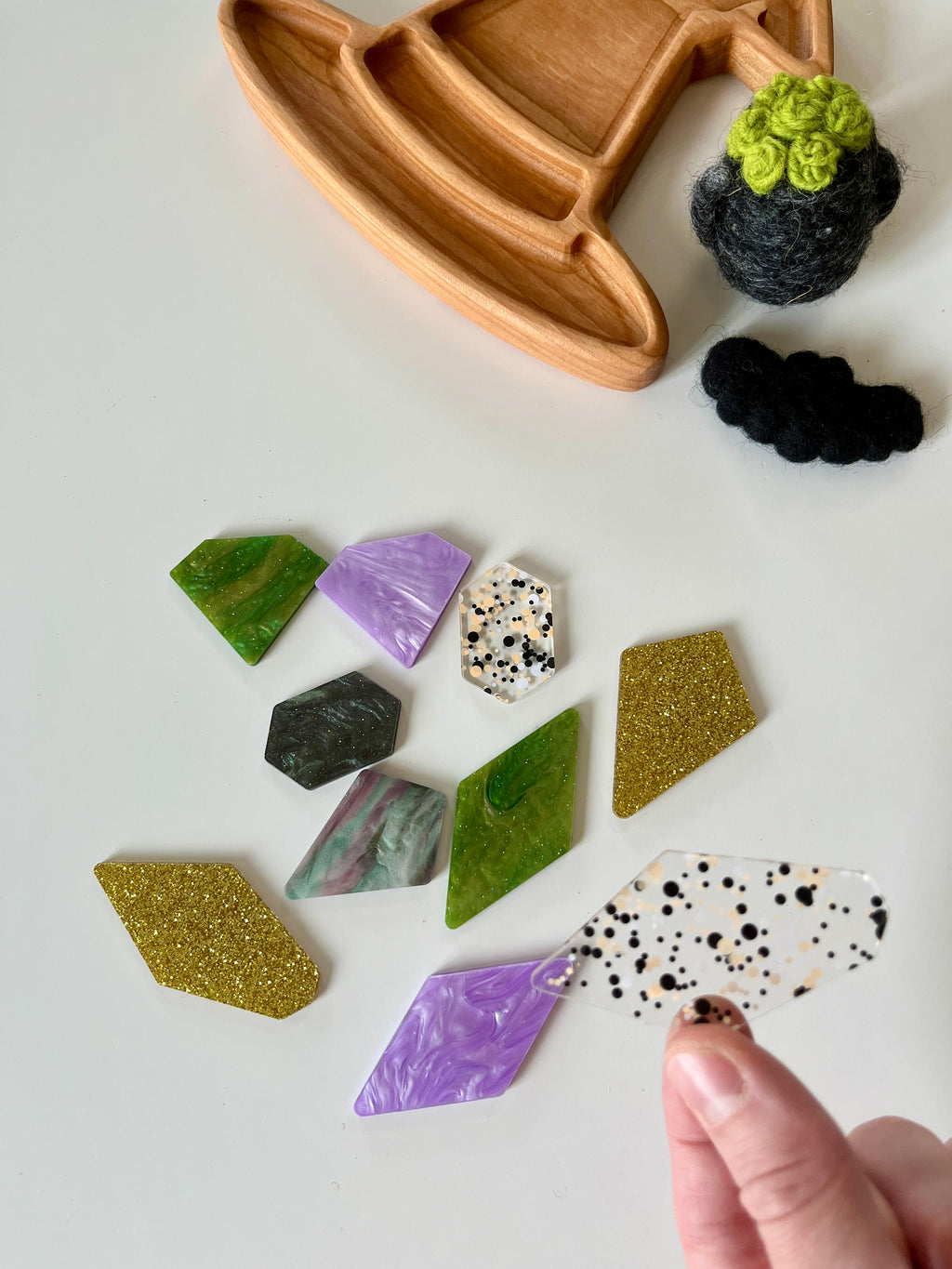 Assorted geometric acrylic shapes in green, purple, gold glitter, marble, and speckled patterns on white surface with wooden tray and felt pieces nearby