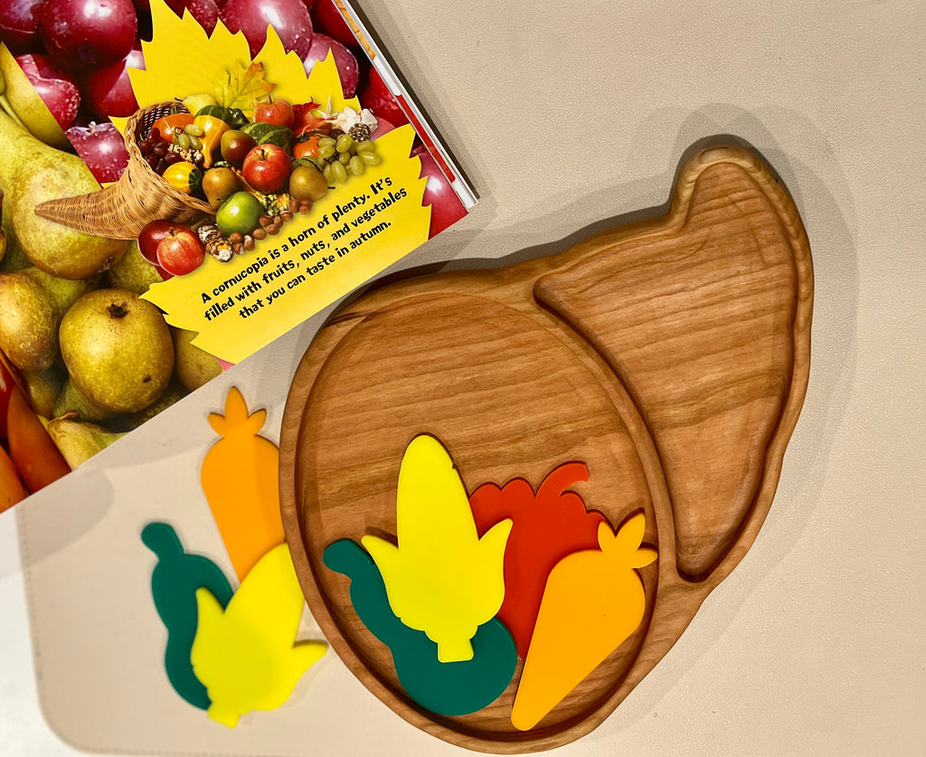Wooden divided plate shaped like a cornucopia with colorful wooden vegetable pieces and a fruit-themed book on beige surface