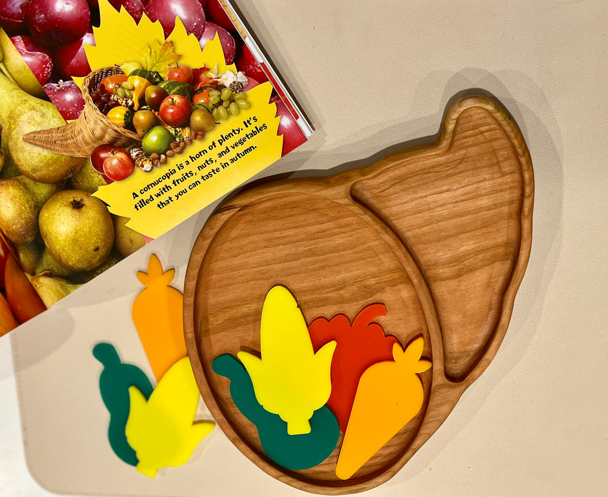 Wooden divided plate shaped like a cornucopia with colorful wooden vegetable pieces and a fruit-themed book on beige surface