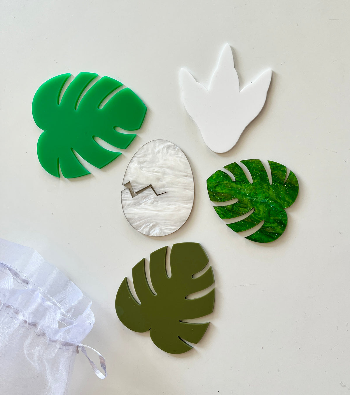 Five various green and white acrylic shapes including tropical leaves and a cracked eggshell on white surface next to sheer fabric pouch