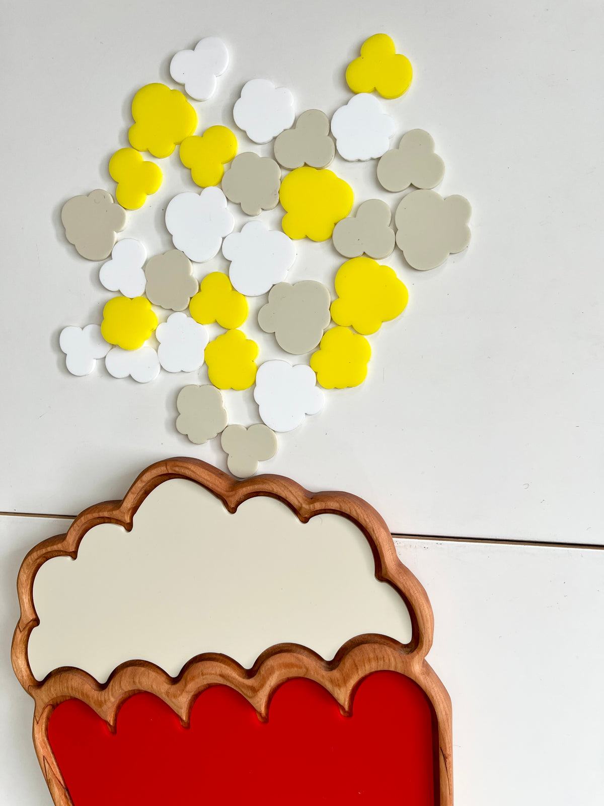 Wooden tray shaped like a popcorn container with red base and scattered yellow, white, and beige clover-shaped tokens on white surface