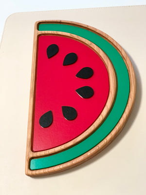 Wooden sensory tray shaped like a watermelon slice with red and green painted sections and six black seeds