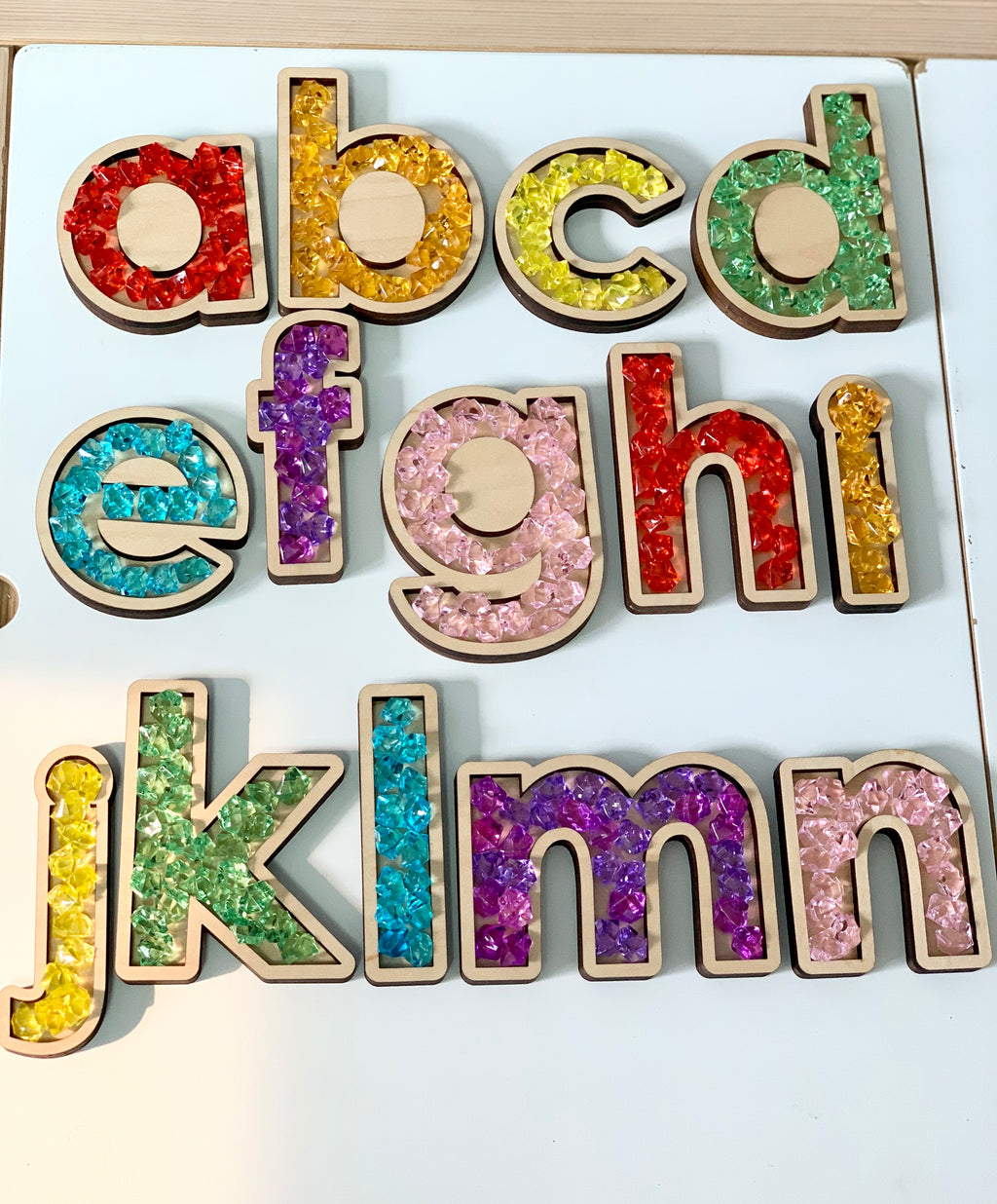 Laser cut wooden alphabet trays filled with colorful translucent sensory beads arranged on white surface