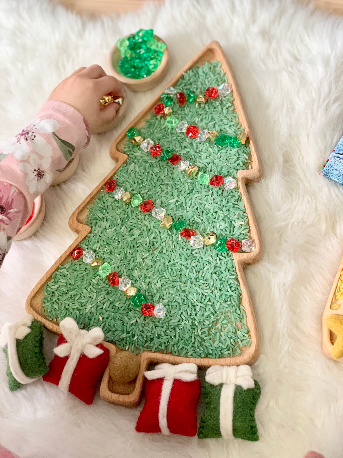 Large wooden tree-shaped sensory tray filled with green rice and translucent beads with felt gift decorations on fur background