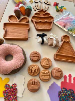 Wooden trays shaped like cupcake, gift box, party hat, carved stamps, felt donuts, and colorful crafting supplies on table