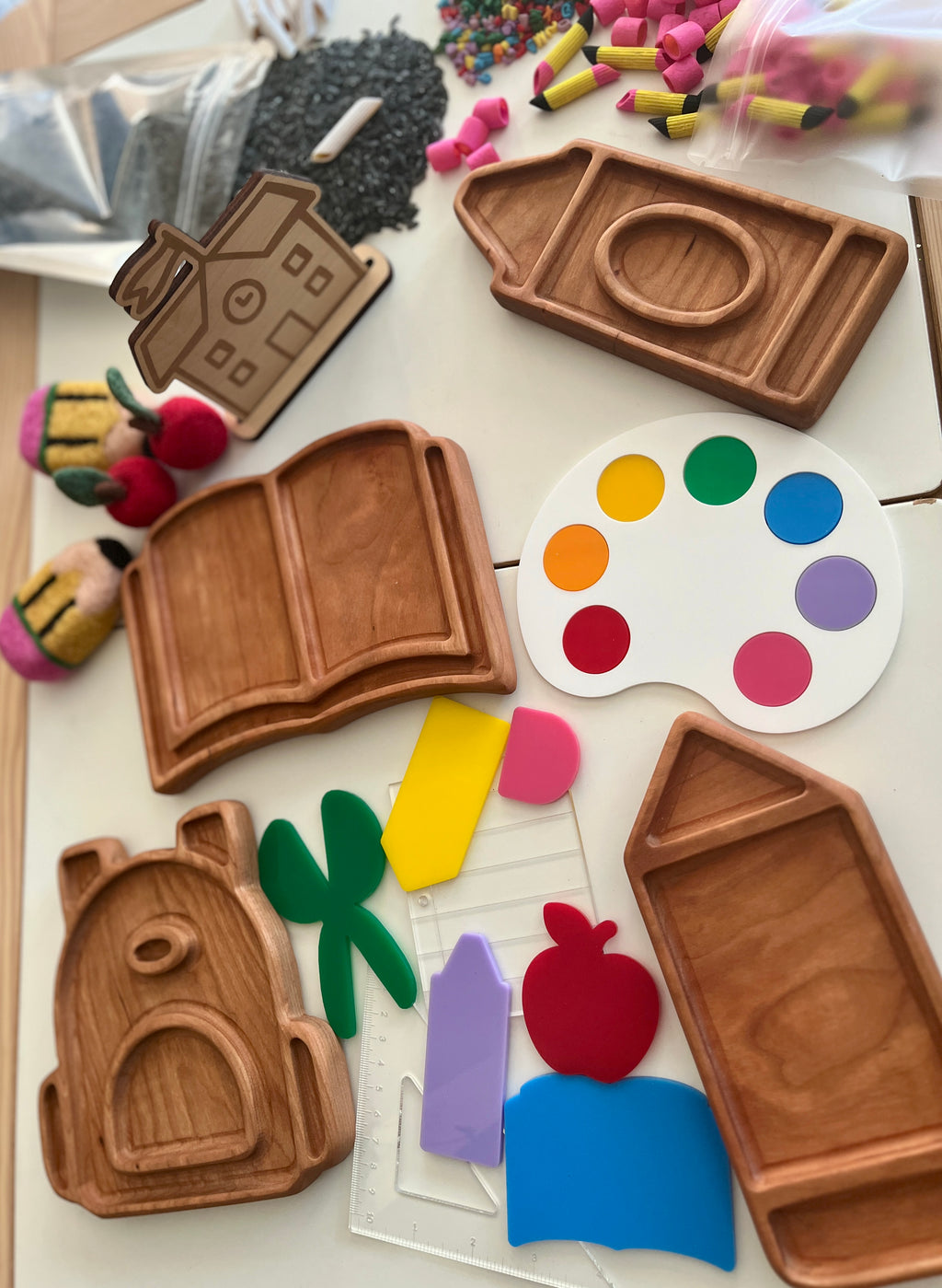 Wooden trays shaped like camera, book, house, and backpack alongside colorful craft pieces and acrylic shapes on table