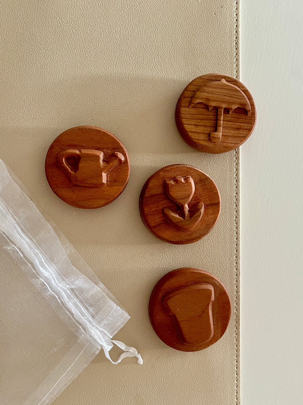 Four round wooden tokens with carved gardening icons and a sheer white drawstring pouch on a beige surface