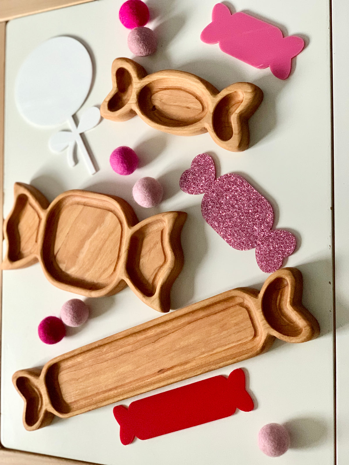 Three wooden candy-shaped trays with pink and red felt balls and glittery and solid candy cutouts on white surface