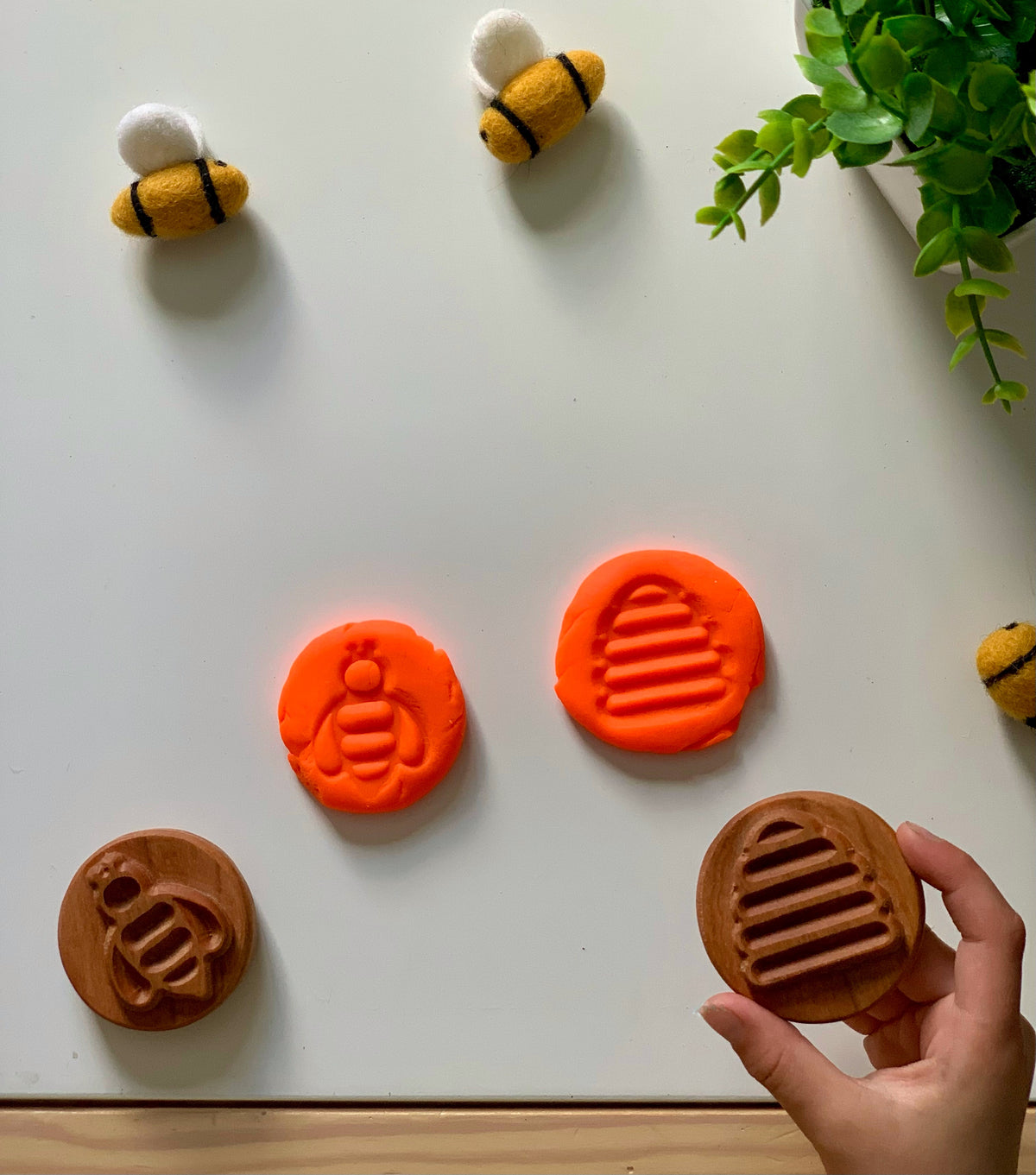Hand holding round wooden stamp with beehive design, matching orange clay impressions, felted bee toys, and green plant on white surface