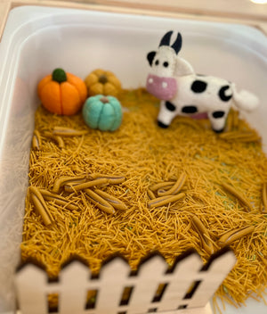 Small white container with dried pasta as ground, felt pumpkins, and a plush cow standing inside a wooden fence