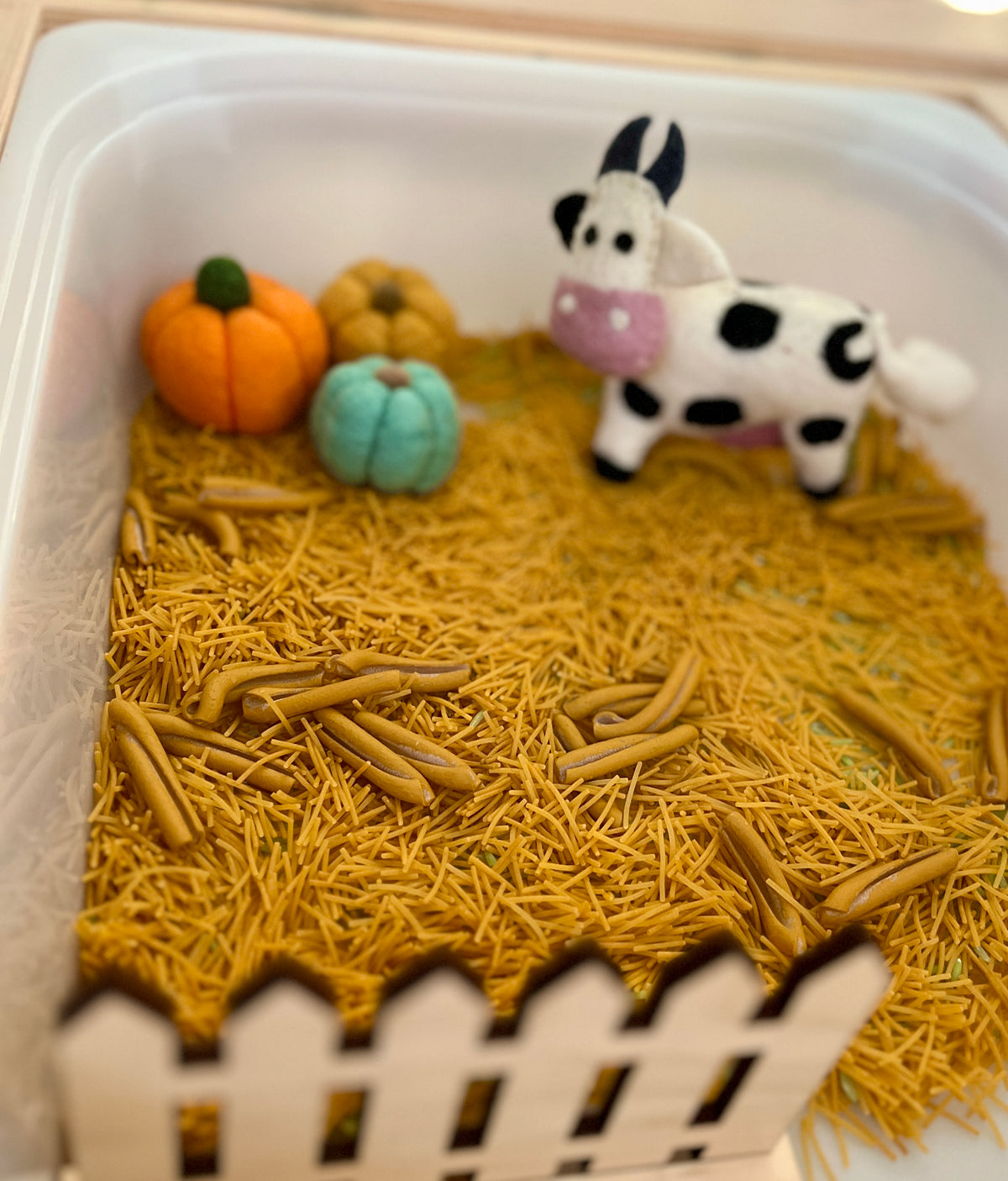 Small white container with dried pasta as ground, felt pumpkins, and a plush cow standing inside a wooden fence
