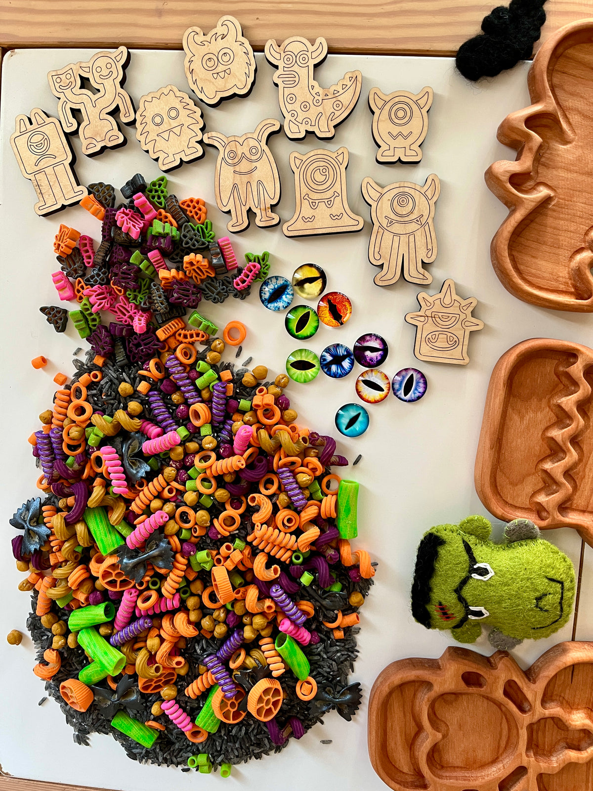 Colorful Halloween-themed pasta and black beans with wooden monster tokens, decorative monster eyes, and pumpkin-shaped trays on white surface