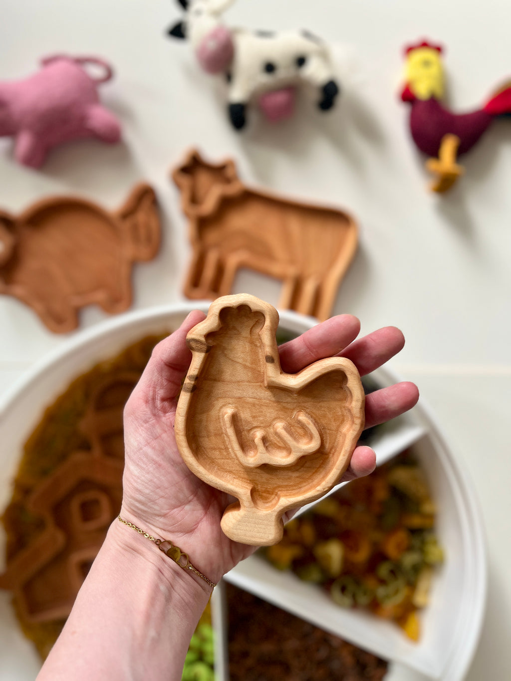 Hand holding small wooden tray shaped like a chicken with three other farm animal trays and felt toys in background