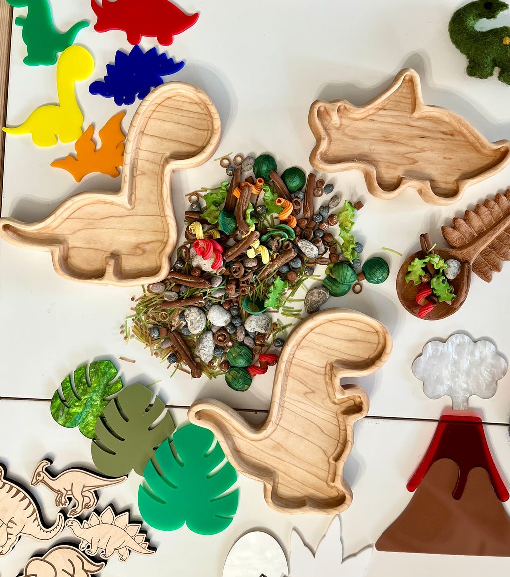 Wooden dinosaur-shaped trays surrounded by colorful dinosaur cutouts, green leaves, natural materials, and a volcano toy display.