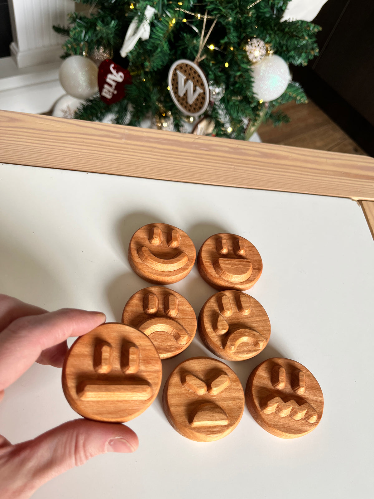 Hand holding round wooden stamp with neutral expression among other stamps with different faces on white table near Christmas tree