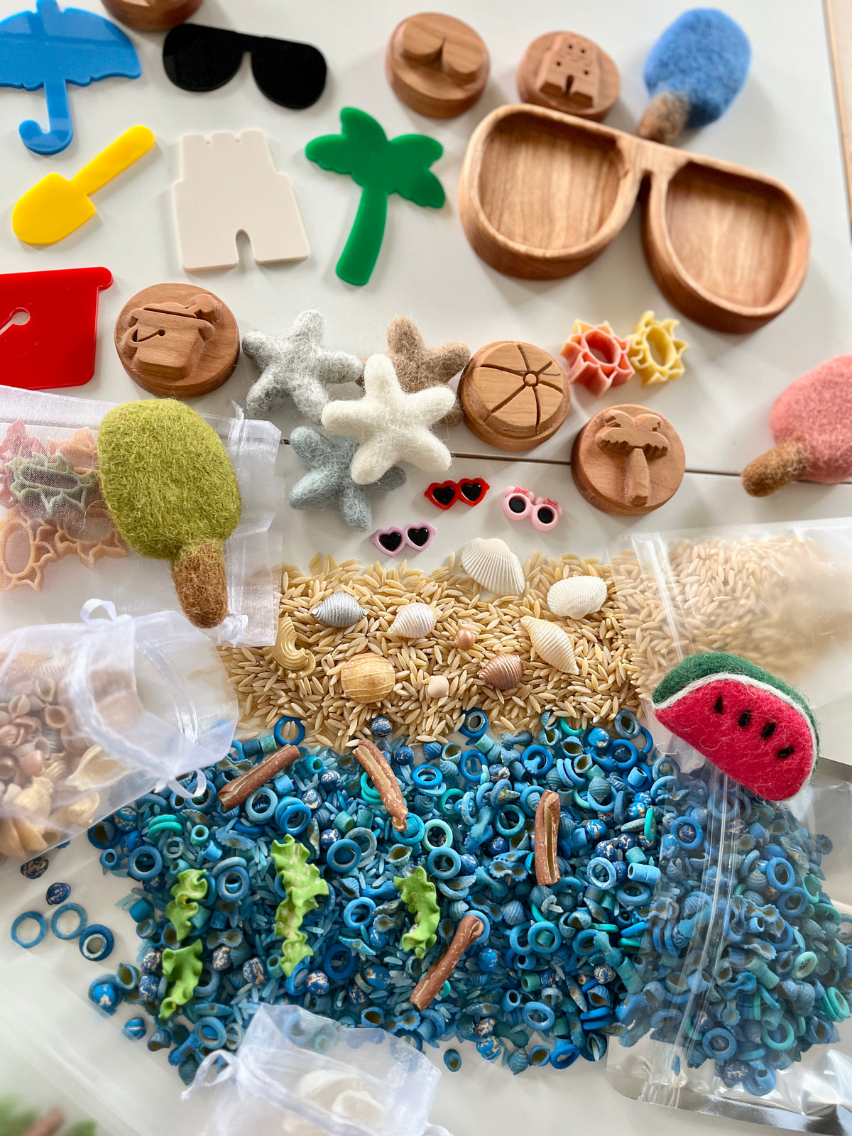 Assorted wooden, felt, and plastic shapes with natural pasta, shells, and blue ring beads arranged on a white surface
