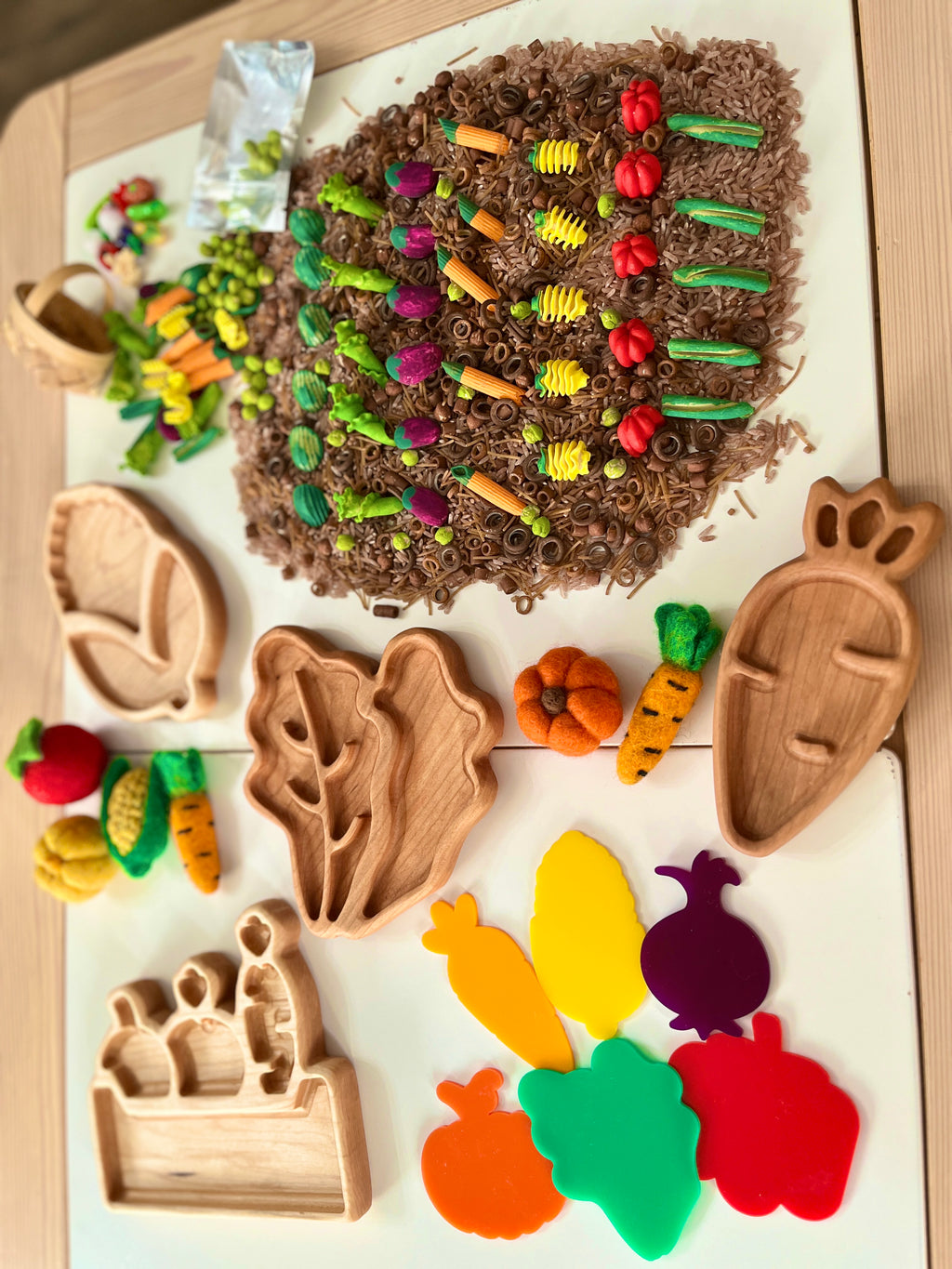 Assorted miniature felt vegetables and wooden trays arranged with brown rice and chocolate curls on a white surface