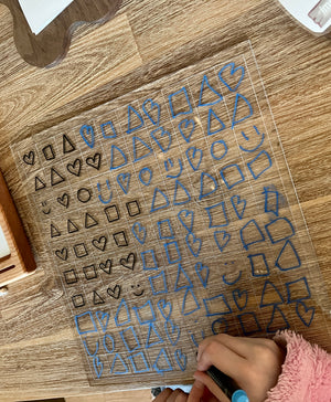 Dry Erasable Blank Open Ended 100 Frame Hundred Board on wooden surface with blue and black hand-drawn shapes including hearts, squares, triangles, and smiley faces, handcrafted homeschool education resources.