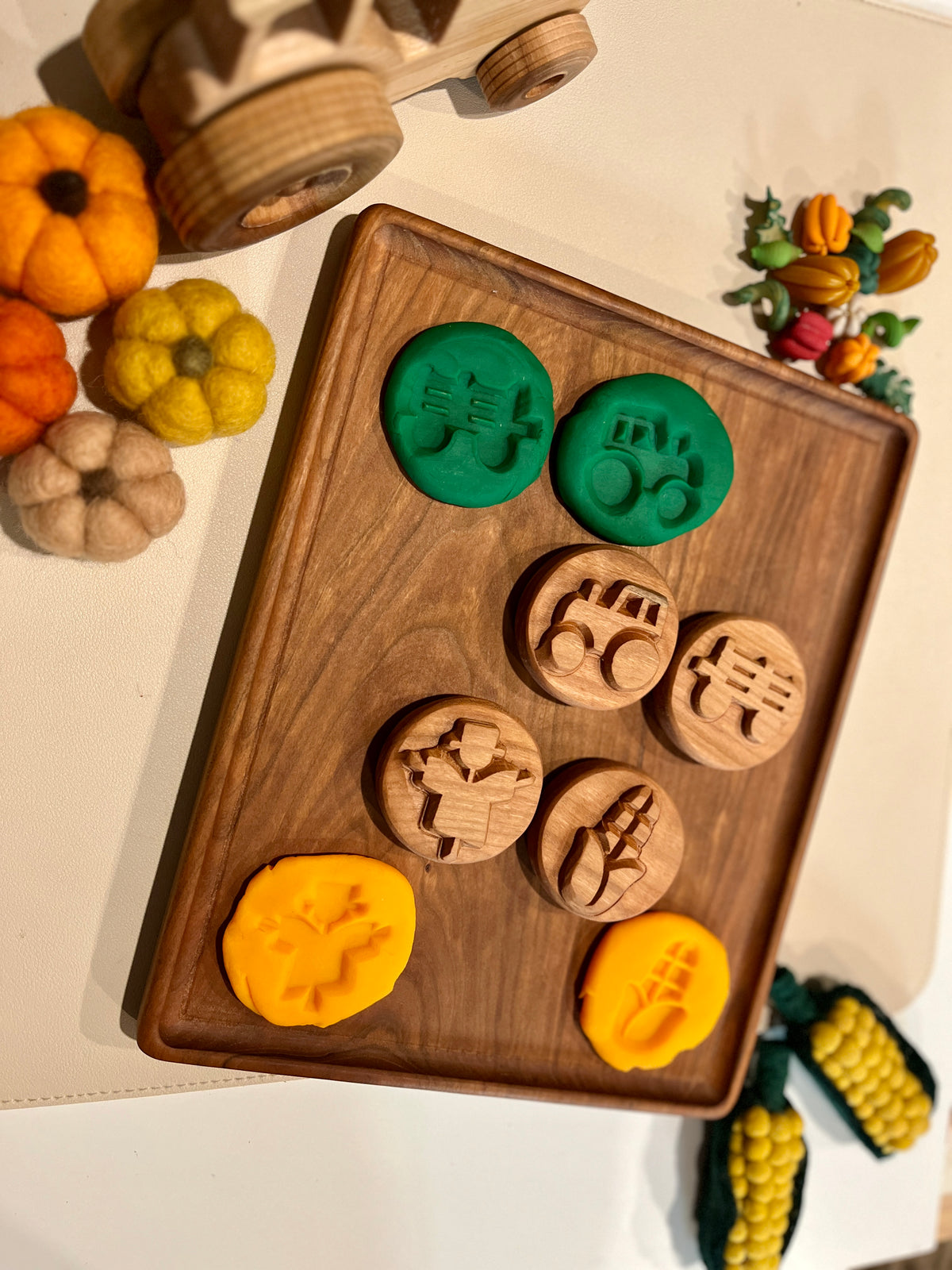 Wooden playdough stamps with green and yellow dough impressions on a wooden tray surrounded by felt pumpkins and corn decorations