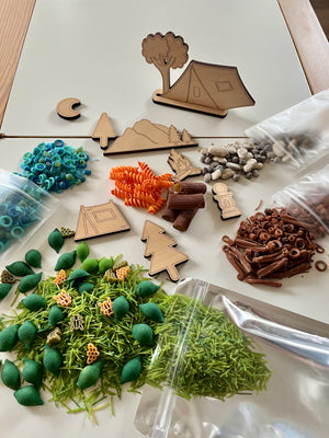 Wooden camping-themed cutouts with colorful pasta shapes, small rocks, and textured play pieces on table surface