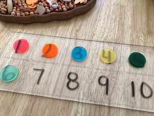 Transparent board with black number stickers from 1 to 10 and colorful translucent circle counters on wooden table