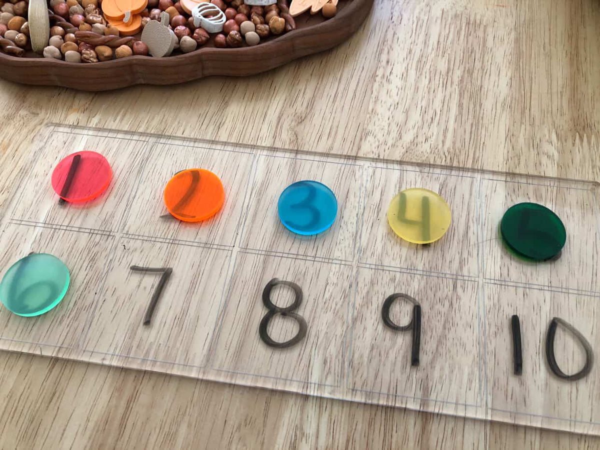 Transparent board with black number stickers from 1 to 10 and colorful translucent circle counters on wooden table