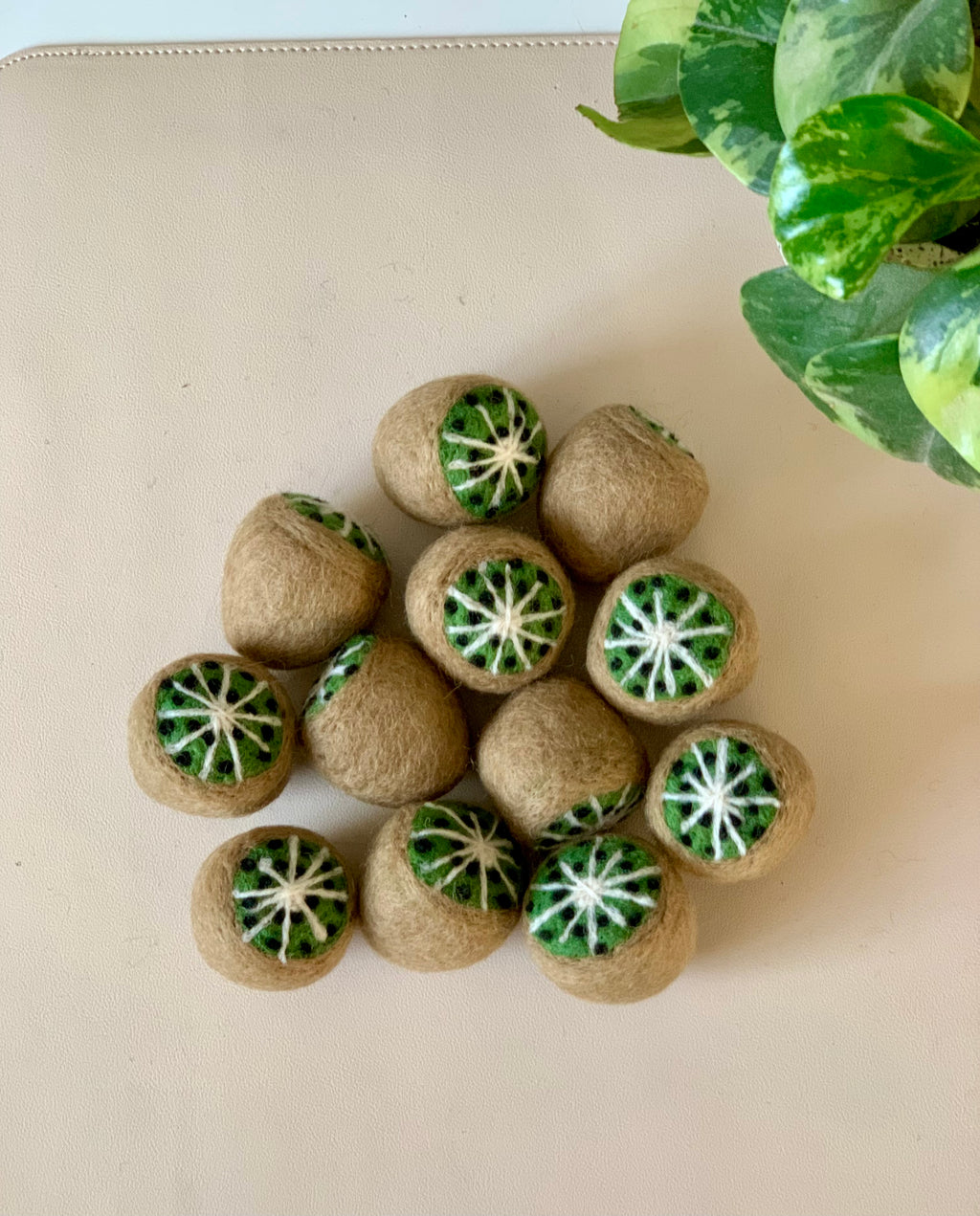 Cluster of small felted brown spheres with green and white kiwi fruit designs on a light surface near variegated green leaves