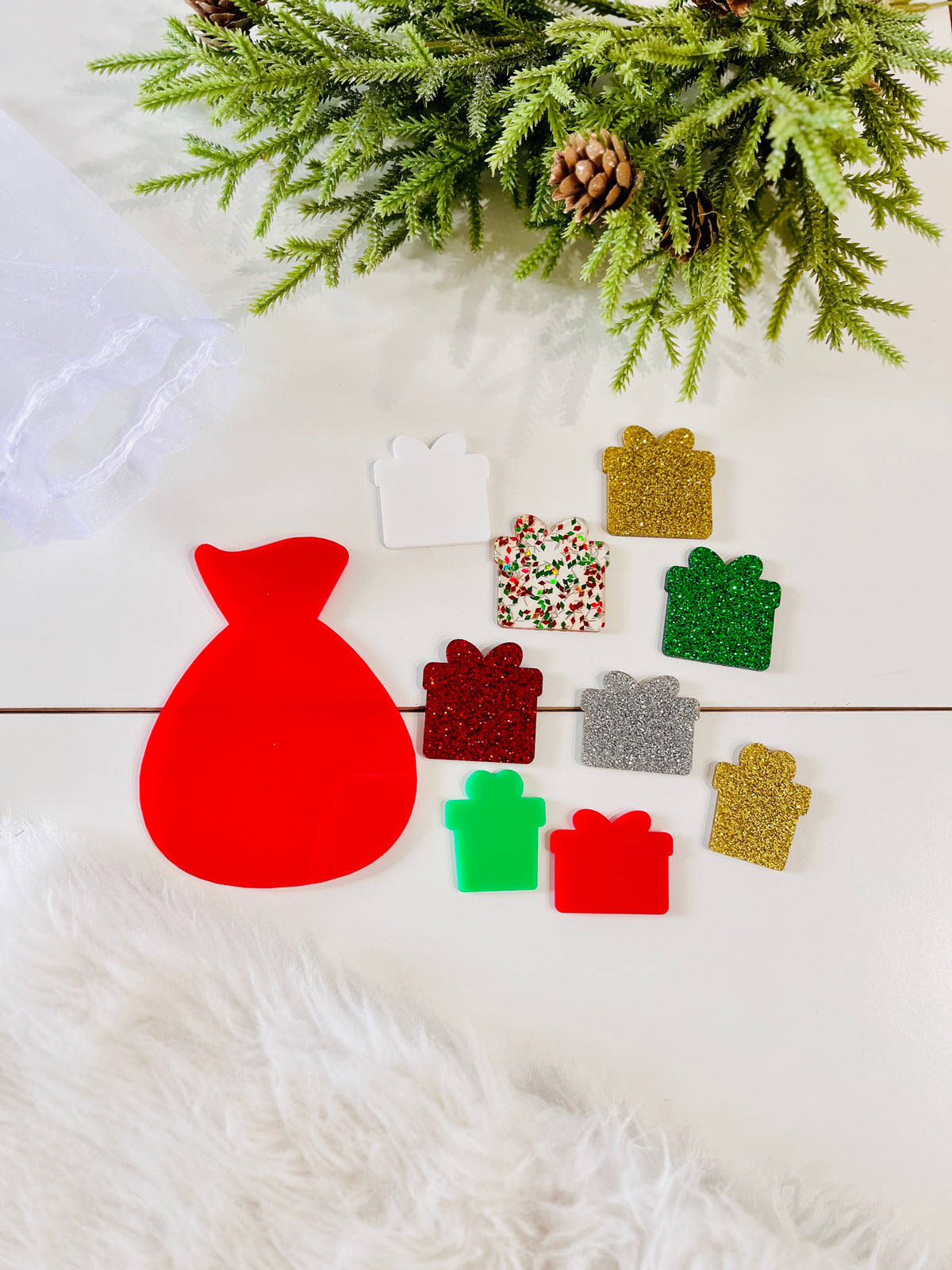 Small colorful gift-shaped tokens arranged near a red sack cutout and pine branch on light surface
