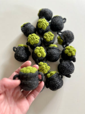 Hand holding one small dark gray felt pot with bright green textured top, with several similar pots scattered on white surface.