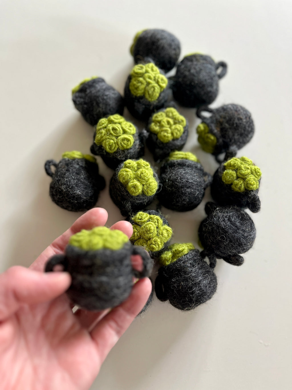 Hand holding one small dark gray felt pot with bright green textured top, with several similar pots scattered on white surface.