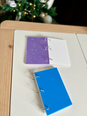Two small three-ring binders with purple and blue plastic covers on a cream and wooden table near a decorated tree