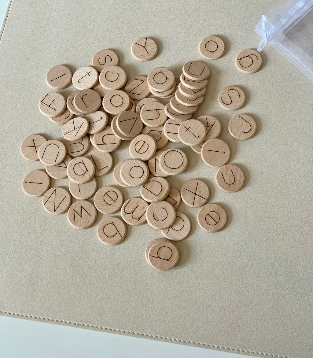 Small natural wooden discs with laser engraved lowercase alphabet letters scattered on beige surface next to mesh bag