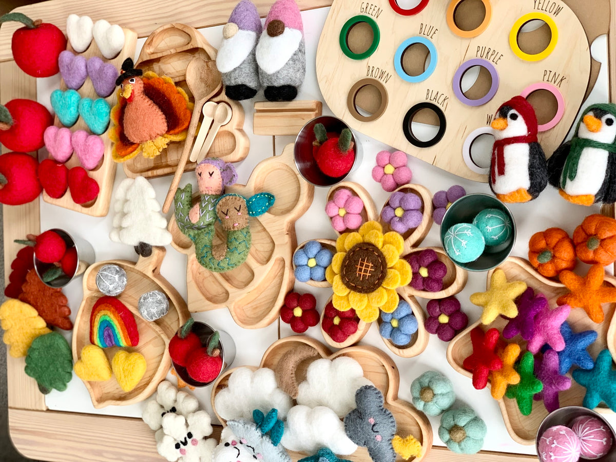 Colorful handcrafted felt flowers, animals, and fruits along with wooden trays and educational color-matching boards arranged on a light wood surface, supporting imaginative homeschool play and sensory learning.