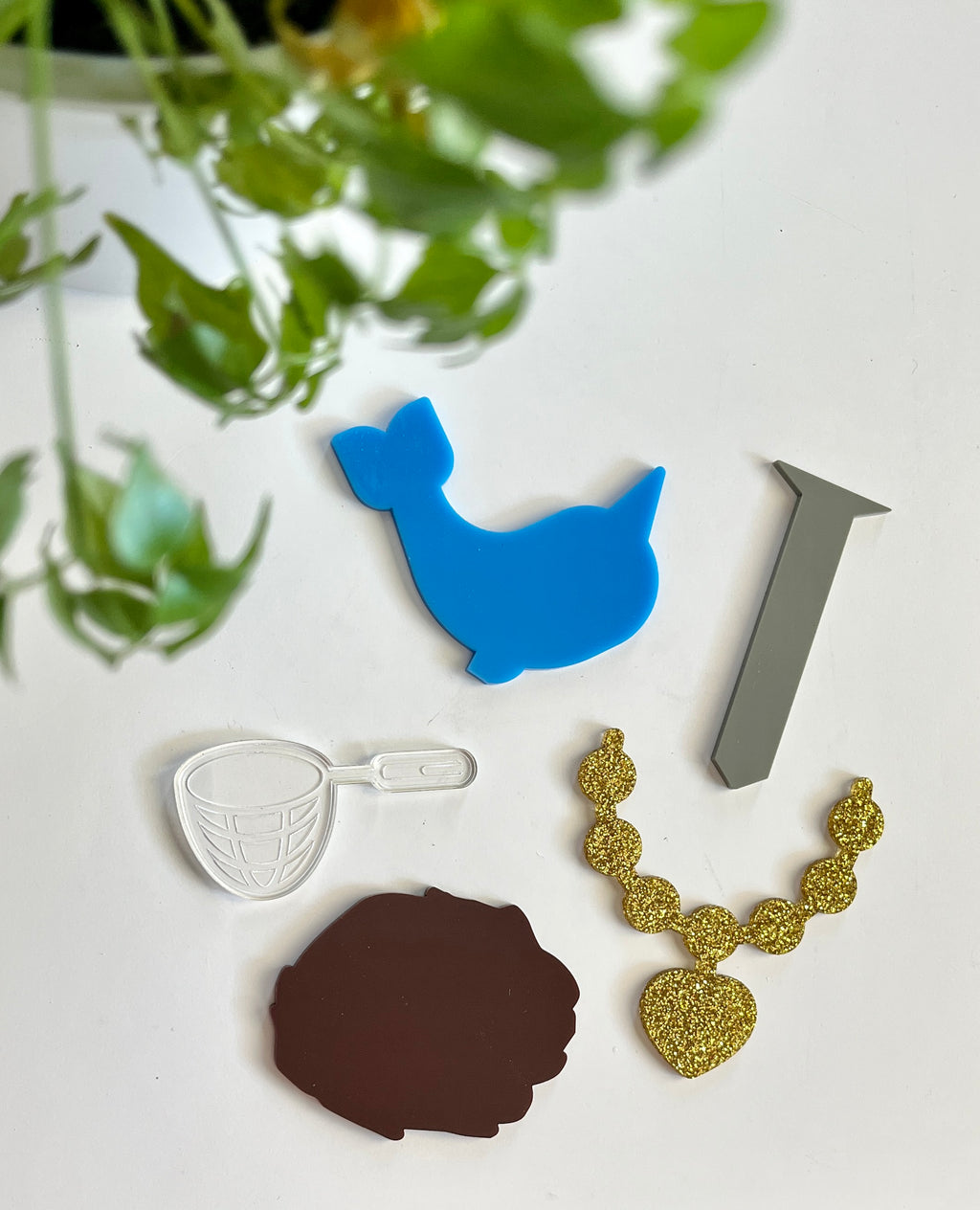 Five colorful acrylic shapes including blue duck, gray nail, transparent sieve, brown blob, and gold glitter necklace on white surface
