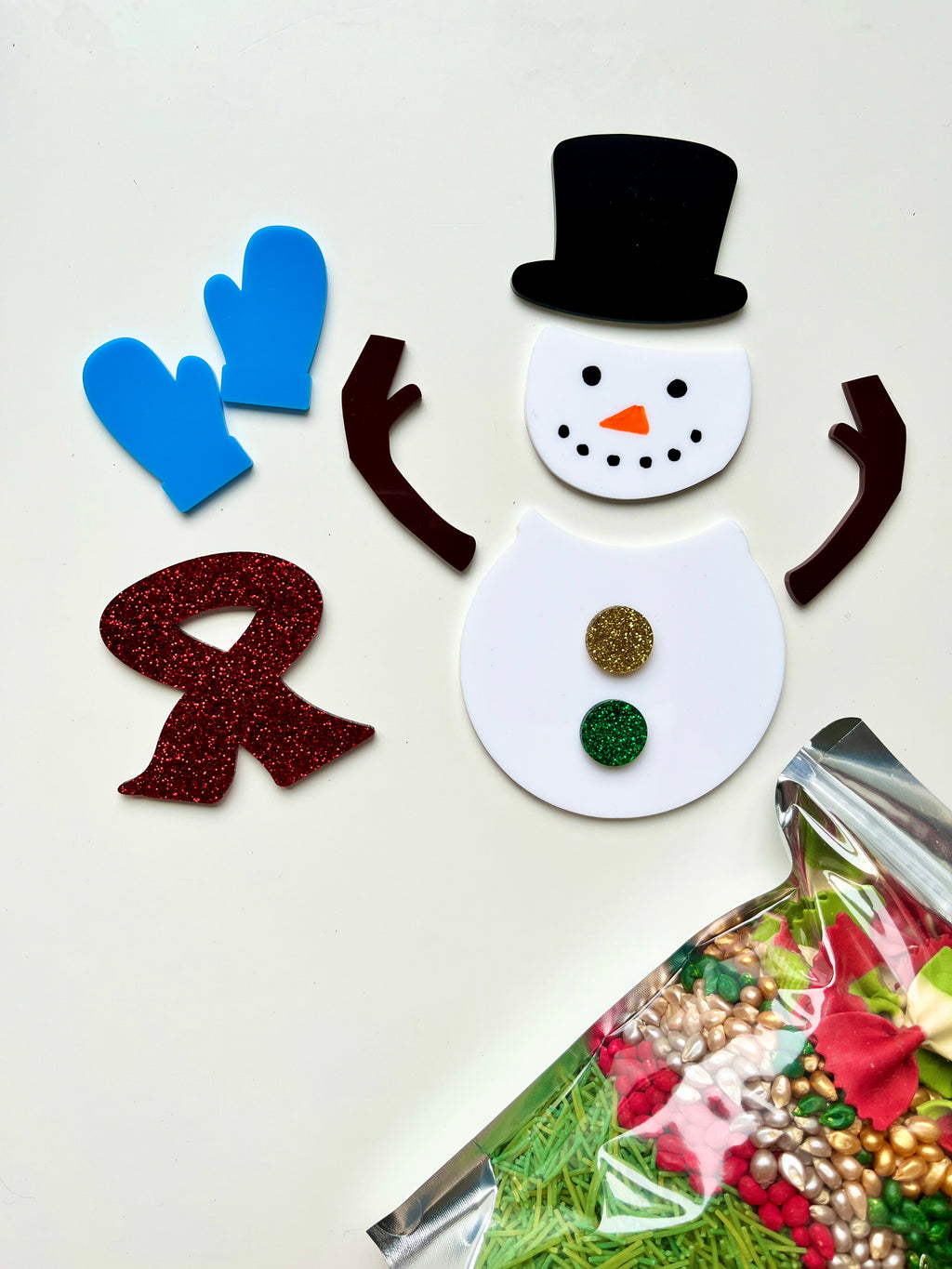 Unassembled snowman craft pieces with black top hat, blue mittens, brown arms, glittery red scarf, and colorful buttons on white background