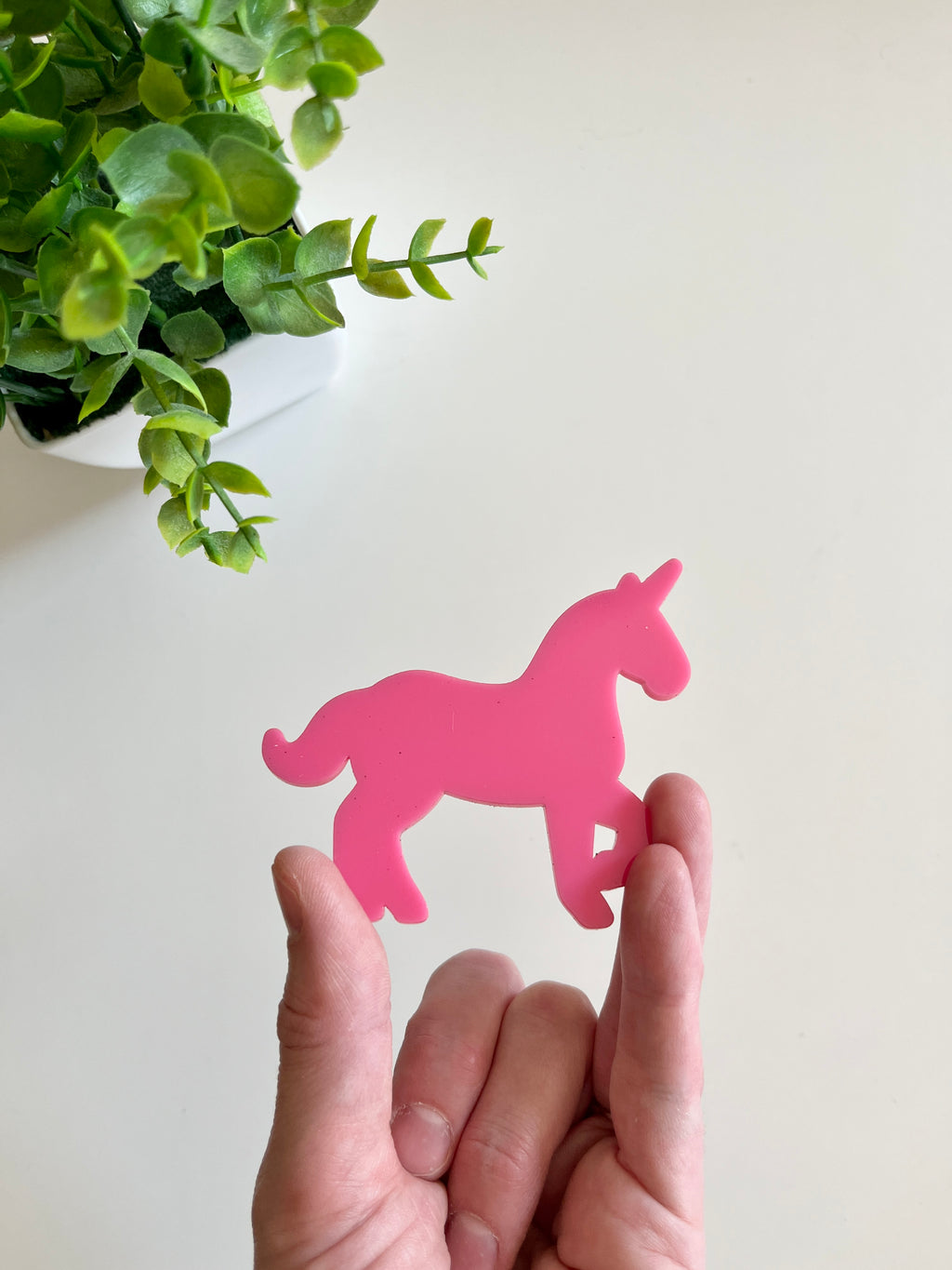 Fingers holding a small bright pink unicorn-shaped object with a green leafy plant in a white pot nearby