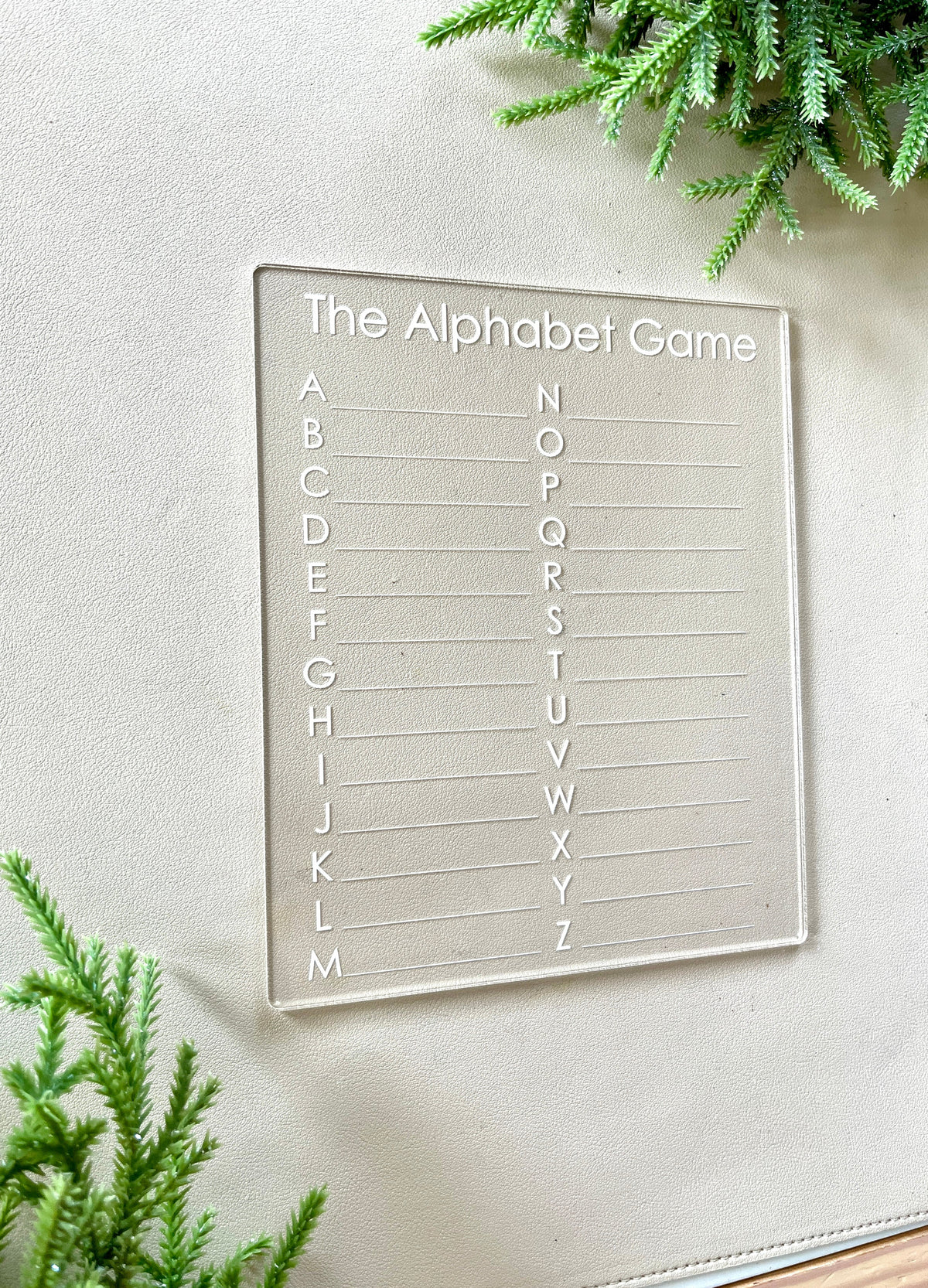 Clear acrylic game board engraved with the alphabet from A to Z and labeled "The Alphabet Game" on a light surface with greenery