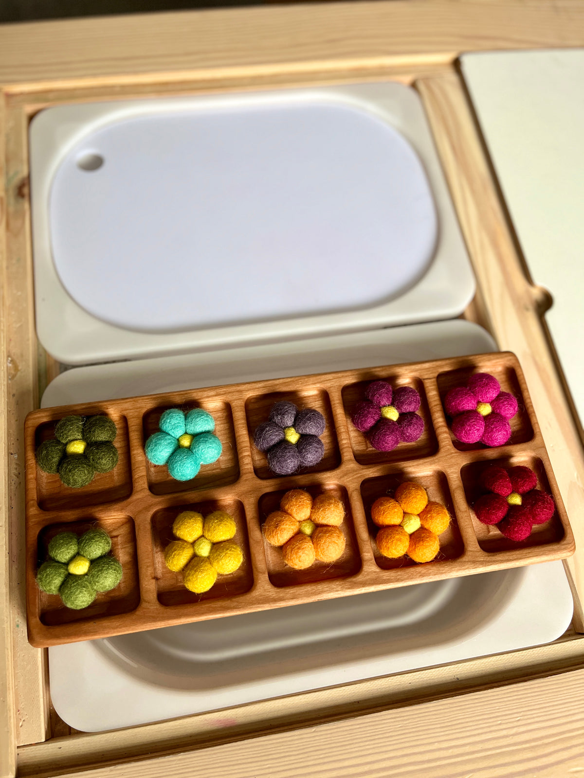Wooden tray with ten compartments each holding a small felt flower in various bright colors on a wooden surface