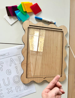 Wooden-framed board with clear acrylic sheet held above, colorful magnetic squares, dry erase marker, and alphabet practice worksheet nearby