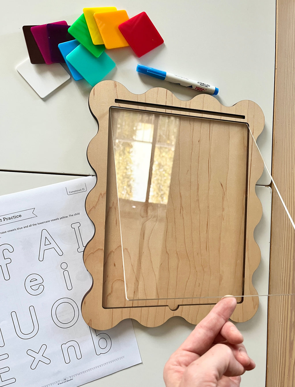 Wooden-framed board with clear acrylic sheet held above, colorful magnetic squares, dry erase marker, and alphabet practice worksheet nearby