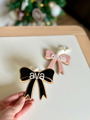 Hand holding black wooden bow decoration with name "ava" and pink bow with name "olivia" blurred on white table